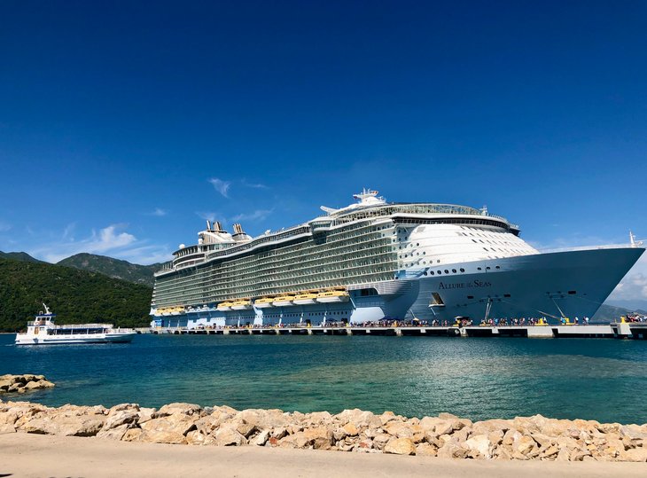 Ein großes weißes Kreuzfahrtschiff dominiert das Bild. Davor ist ein Hafen und dahinter eine Hügellandschaft.