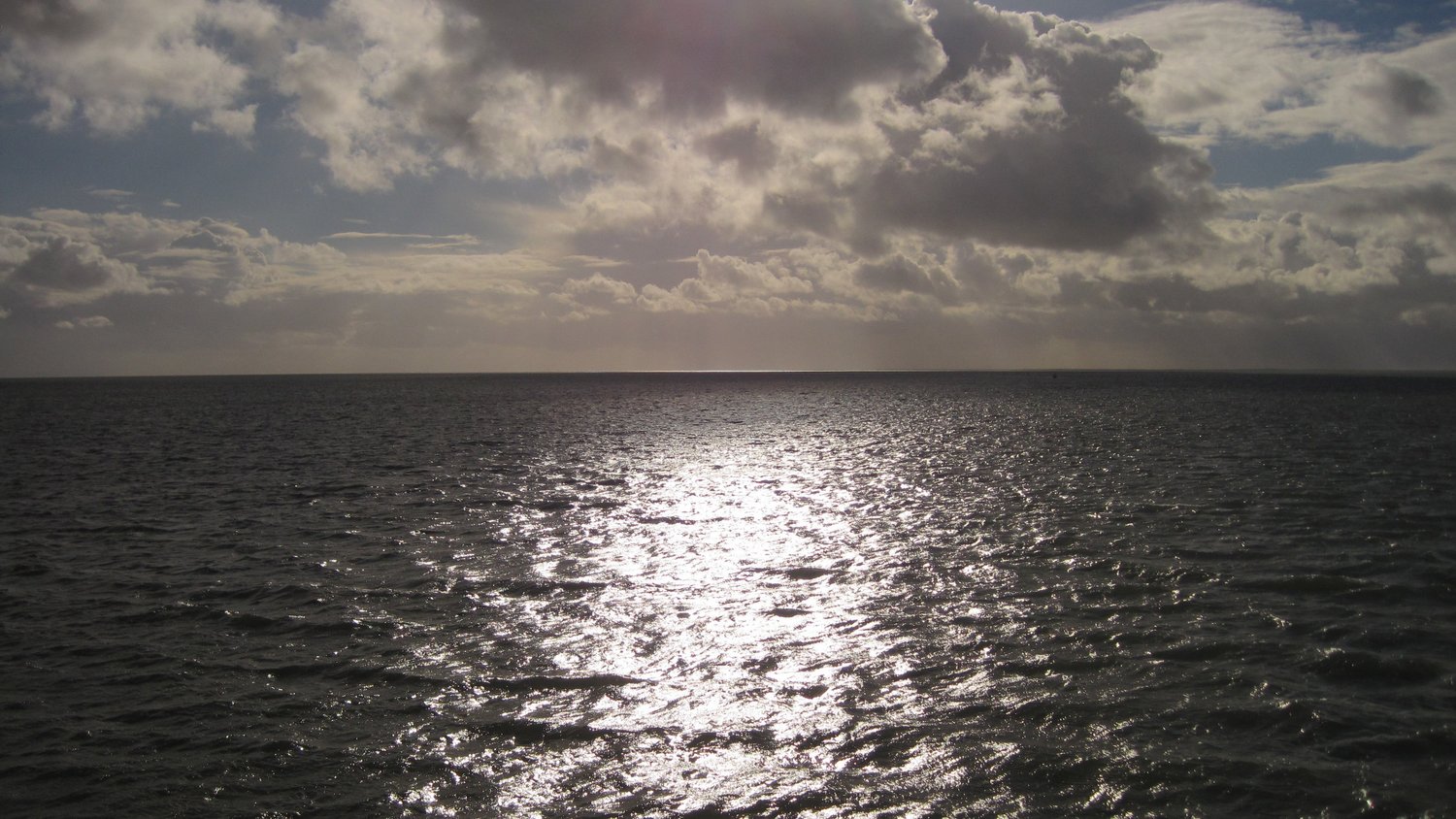 Das Bild zeigt die Wasserfläche der Nordsee, in der sich die Sonne spiegelt. Es sind keine Wellen zu sehen, das Wasser ist glatt. Am Himmel sind graue Wolken, die aber Lücken haben, durch die Sonnenstrahlen auf das Wasser fallen.