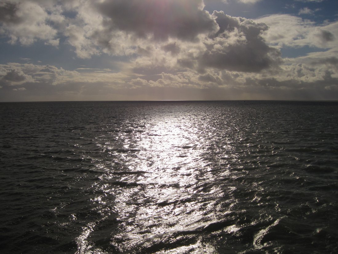 Das Bild zeigt die Wasserfläche der Nordsee, in der sich die Sonne spiegelt. Es sind keine Wellen zu sehen, das Wasser ist glatt. Am Himmel sind graue Wolken, die aber Lücken haben, durch die Sonnenstrahlen auf das Wasser fallen.